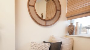 A wooden chest with pillows, a round mirror, and a window with blinds.