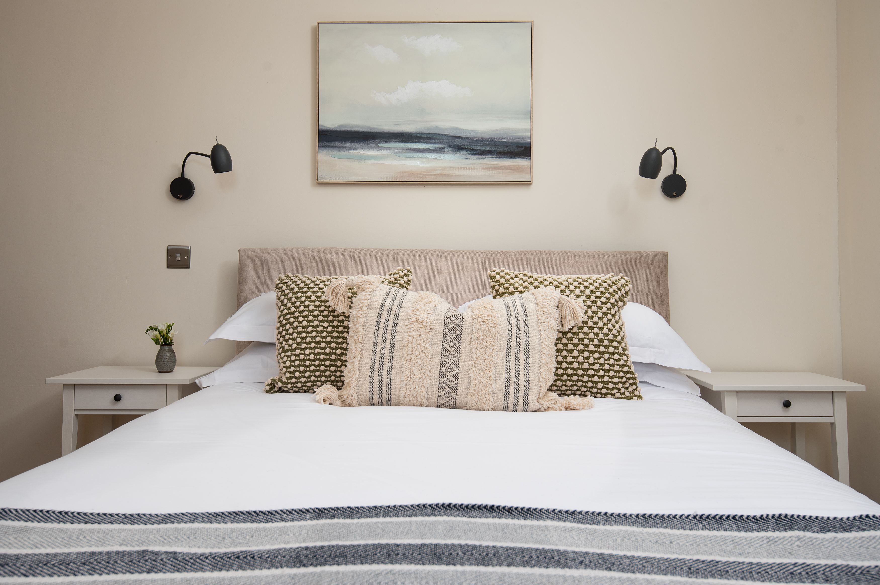 A neatly arranged bedroom with a large bed, two bedside tables, a wall-mounted painting, and two wall sconces.