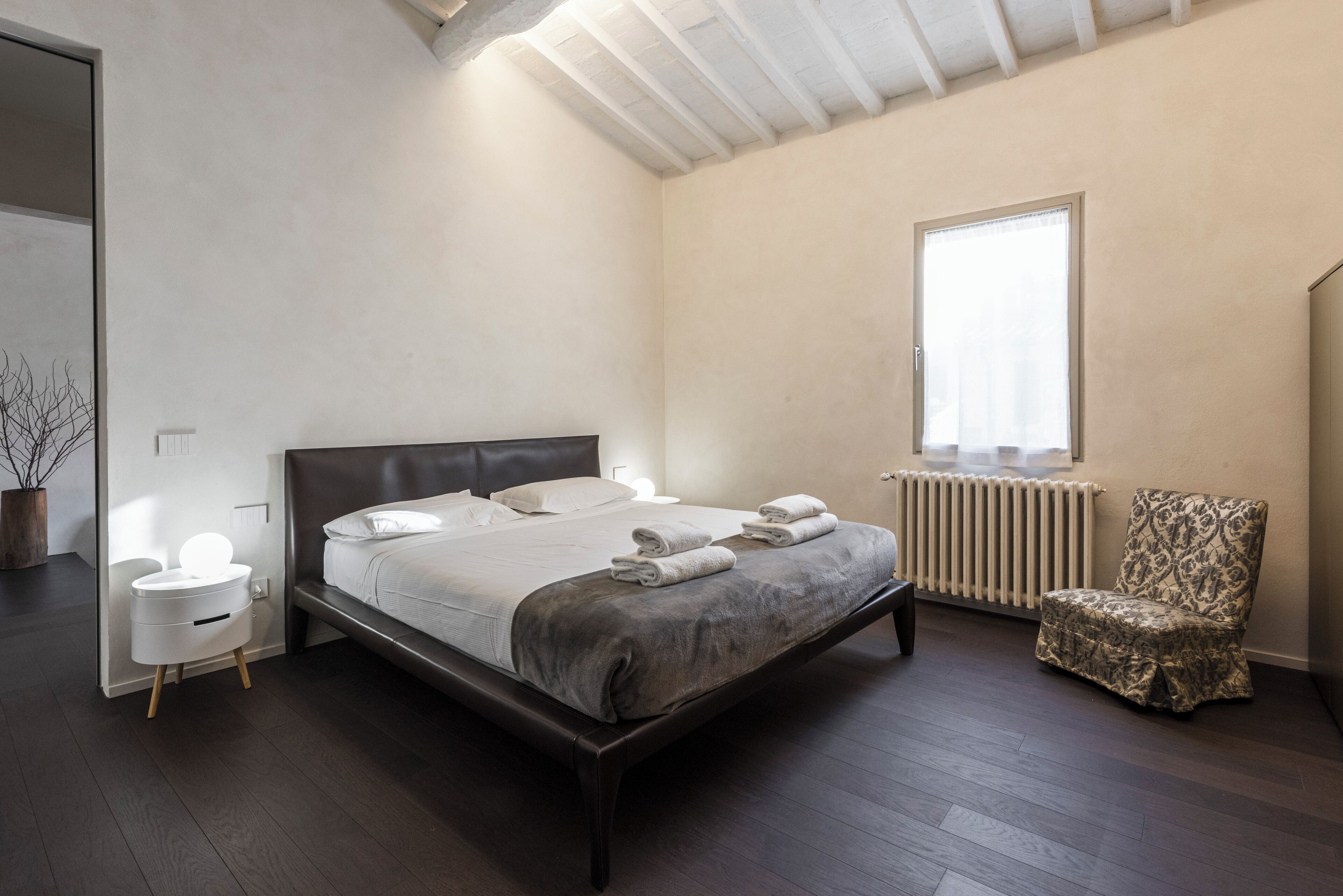Un dormitorio con una cama grande, mesitas de noche, una silla, un radiador y una ventana.