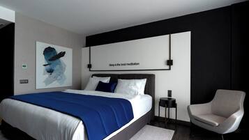 A modern bedroom with a large bed, a side table, a chair, and a wall-mounted artwork.