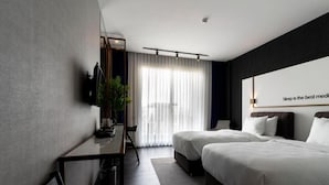 Ett modernt hotellrum med två sängar, ett skrivbord och ett stort fönster med tunna, genomskinliga gardiner.