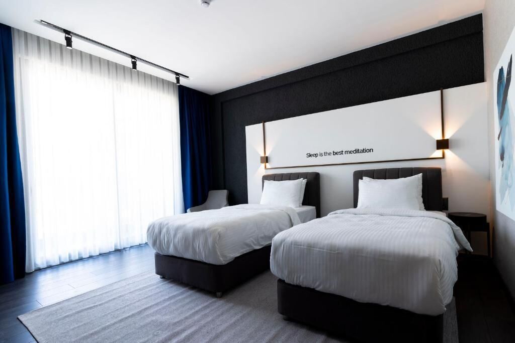 A hotel room with two beds, a large window with curtains, and a wall-mounted sign that reads "Sleep is the best meditation."