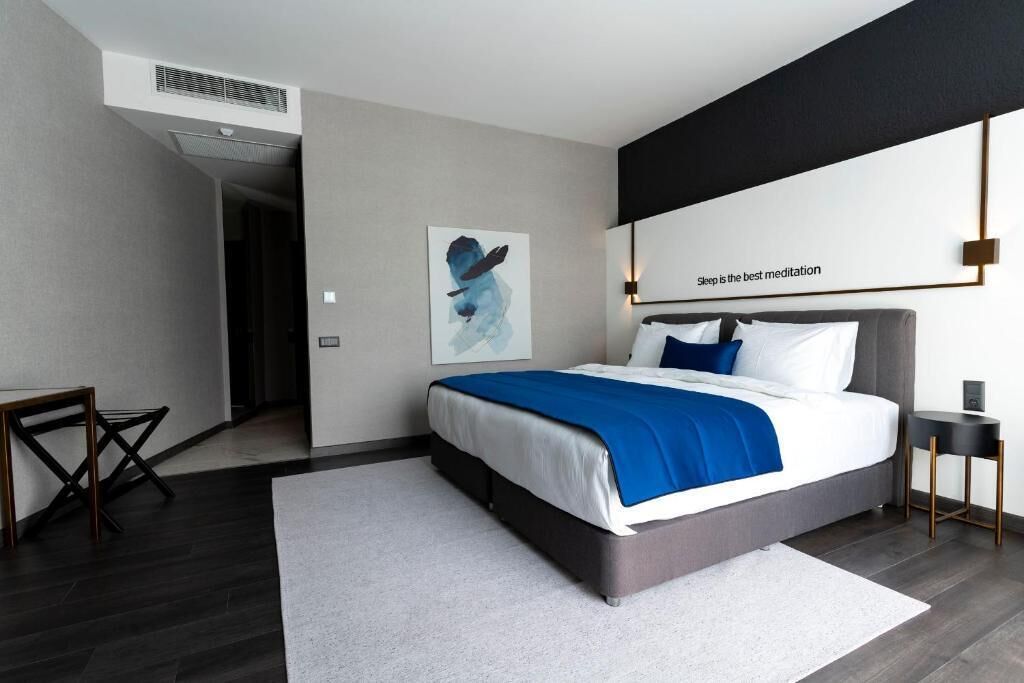 Une chambre moderne avec un grand lit, une table de chevet, une lampe fixée au mur et une œuvre d’art encadrée.