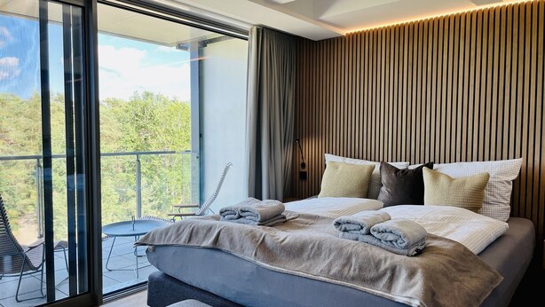 A modern bedroom with a large bed, a view through a sliding glass door, and a wooden slatted wall. - "Purbaltic" maisonette apartment with lake view and sauna, completed in 2022 (Prora)