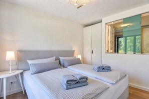 A hotel room with two beds, a bedside table with a lamp, a window with a view of greenery, and a mirror on the wall.