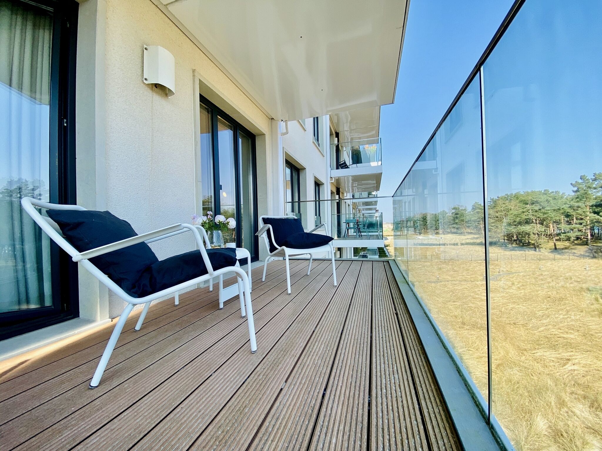A balcony with two white chairs and a table, overlooking a grassy area with trees.