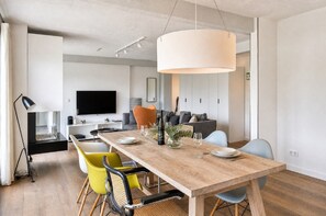 A modern dining area with a wooden table, yellow and blue chairs, a large pendant light, and a television in the background.