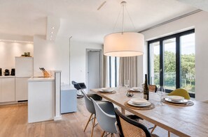 A modern kitchen with white cabinetry, a dining area with a wooden table, and large windows offering a view of greenery. - Meerloft 13 at Prora Düne 7, with a lake view, private sauna, and fireplace (Prora)