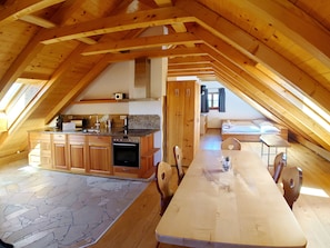 A spacious attic bedroom with a large bed, a wooden dining table, and a kitchen area with wooden cabinets and a stove.