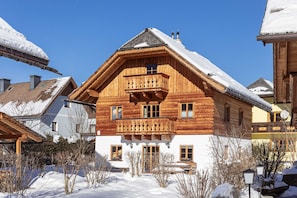 A wooden chalet with a balcony, surrounded by snow-covered trees and other buildings.