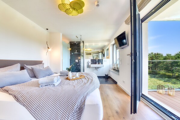 A modern bedroom with a large bed, a television, a bathroom with a mirror, and a view of greenery outside.