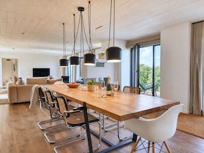 Et moderne spiseområde med et træbord, metalstole, hængende sorte lamper og udsigt til grøn vegetation udenfor.