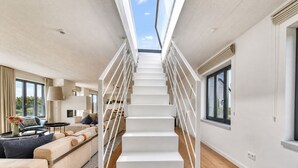Un interior moderno con una escalera blanca central, un conjunto de sofás y un comedor.