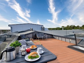 En tagterrasse med et bord dækket til et måltid, inklusive en sandwich, et æble og kaffe, samt en elkedel.