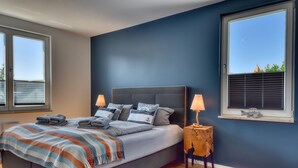 A bedroom with a bed, two bedside lamps, a window with blinds, and a view of the outside.