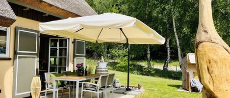 Ein Haus mit Strohdach, einer Terrasse mit Tisch und Stühlen unter einem großen Sonnenschirm.