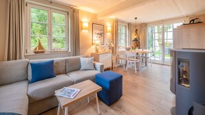 A cozy living room with a sofa, ottoman, and a table with a magazine. There is a dining area with a table and chairs, and a kitchen area with a stove. Large windows provide natural light and views of greenery outside.