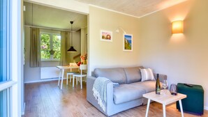 A modern living room with a grey sofa, a small white table, and a green ottoman. There is a dining area with a white table and chairs, and a window with curtains.