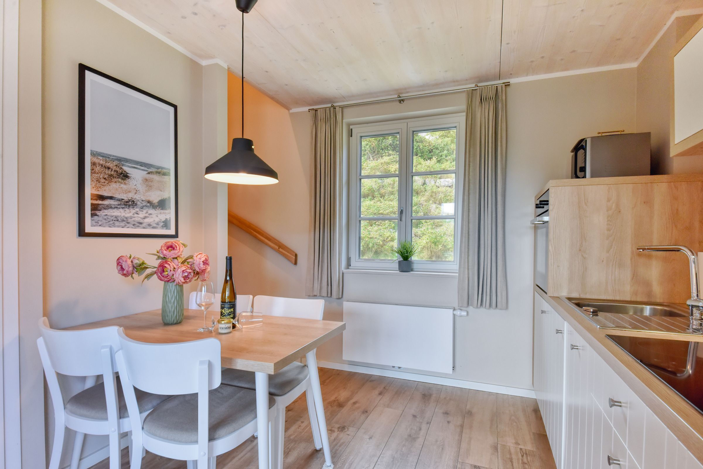 Een moderne keuken met een houten tafel, witte stoelen, een hanglamp, een raam met gordijnen en uitzicht op groen.