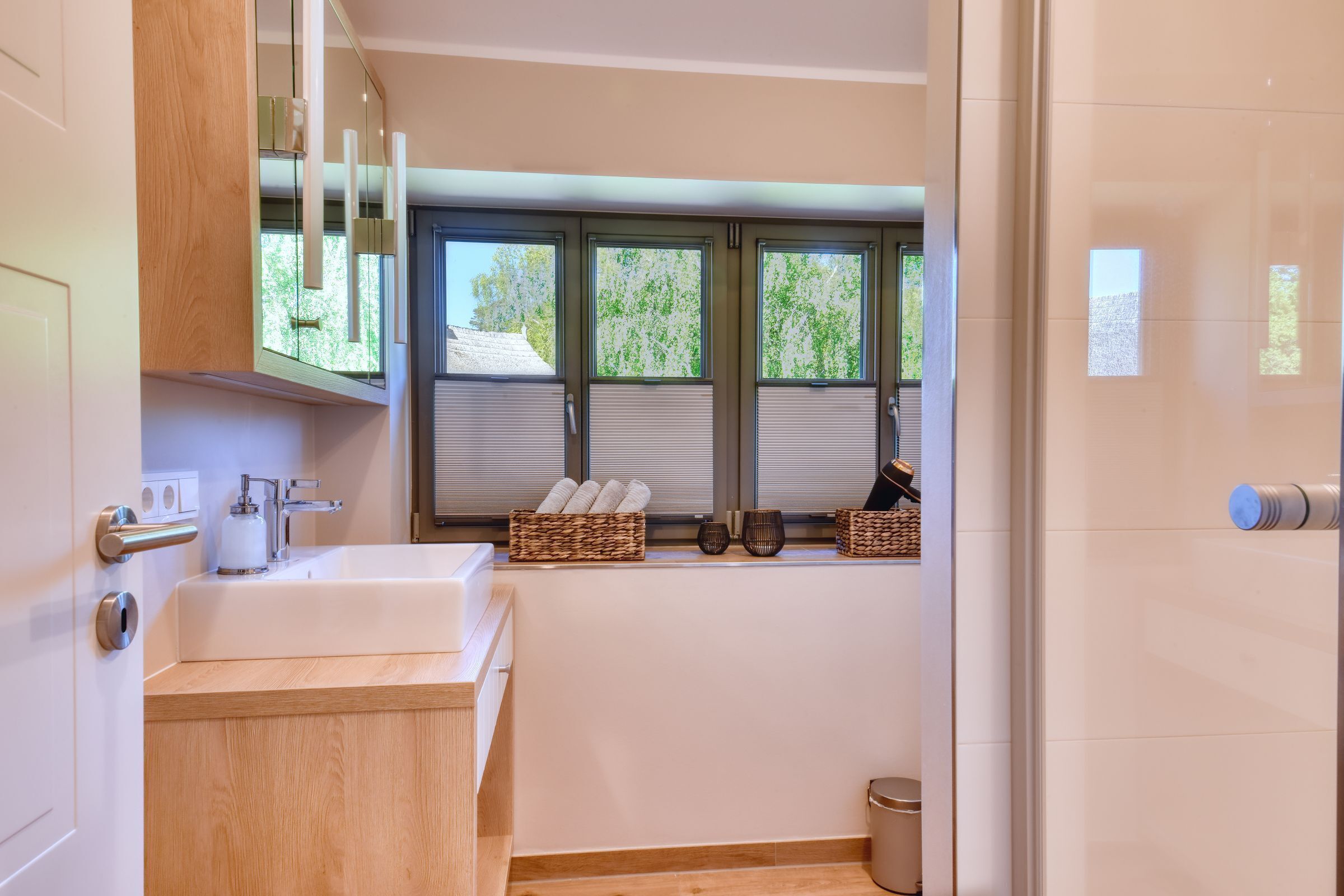 Ein modernes Badezimmer mit einem Holzkosmetikschrank, einem großen Waschbecken und einem Fenster mit Jalousien. Auf dem Fensterbrett stehen Korbwannen und in der Ecke ein Mülleimer.
