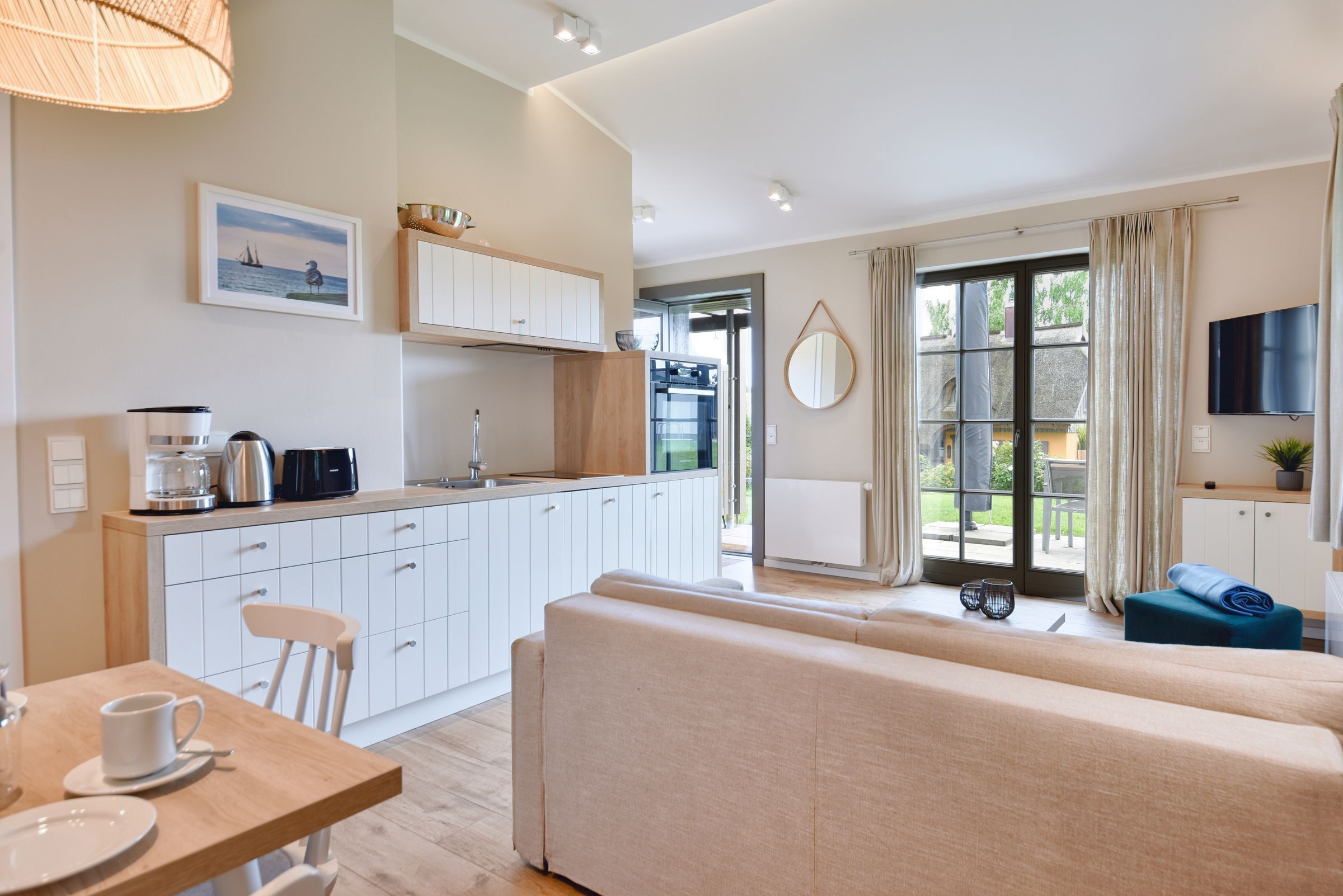 A modern kitchen with white cabinets, a dining table with a cup and saucer, a sofa, and a TV mounted on the wall.