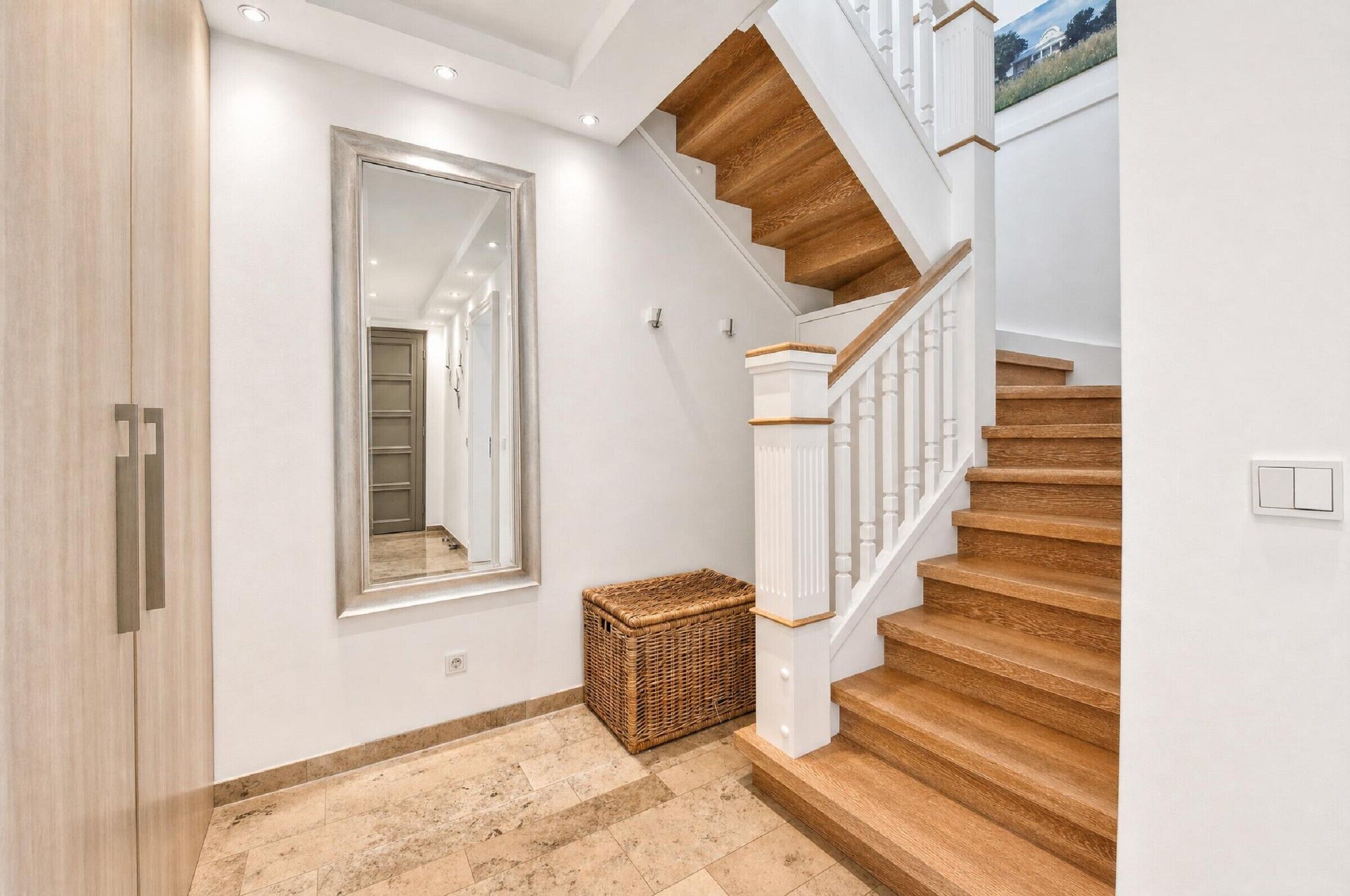 Eine Treppe mit Holzbalken und weißen Geländern, ein großer Spiegel an der Wand und ein Korbwägelchen auf dem Boden.