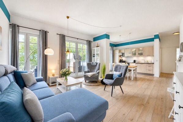 A modern living room with a sofa, chairs, and a coffee table. There is a kitchen area with cabinets and a dining table.