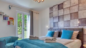 Une chambre à coucher moderne avec un grand lit, un fauteuil bleu, une bibliothèque et une vue sur la verdure par la fenêtre.