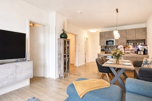 A modern living room with a flat-screen TV, a dining area with a table set for four, and a kitchen with a hanging light fixture.