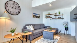 Una sala moderna con un sofá gris, una mesa de centro redonda de madera, una mesita auxiliar con objetos decorativos, un reloj con mapa del mundo y estanterías empotradas con plantas.