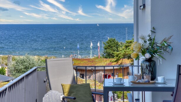 Un balcón con vista al mar, una mesa con tazas y un florero, y una silla con cojín.