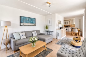 A modern living room with a grey sofa, a wooden coffee table, and a grey armchair. There is a painting on the wall and a potted plant on a small table.