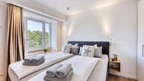 Habitación de hotel con dos camas, una ventana con cortinas, una mesita de noche con lámpara, y un velador con un libro y control remoto.