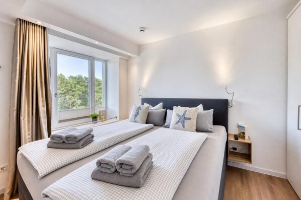 Habitación de hotel con dos camas, una ventana con cortinas, una mesita de noche con lámpara, y un velador con un libro y control remoto.
