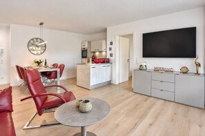 A modern living room with a dining area, a kitchen, and a flat-screen TV mounted on the wall.