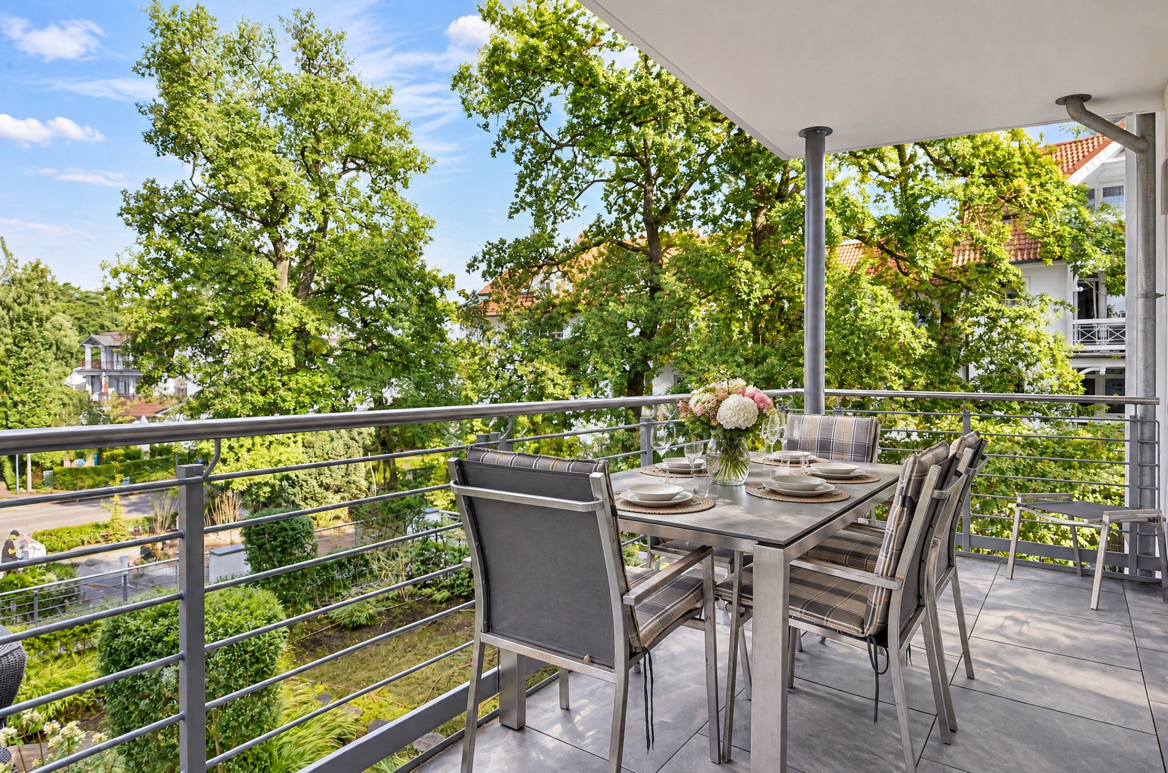 An outdoor dining area with a table set for four, surrounded by chairs, on a balcony with a view of trees and buildings.