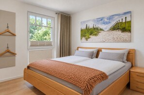 Une chambre avec un lit en bois, une fenêtre avec des rideaux, un tableau représentant une plage et un plancher en bois.