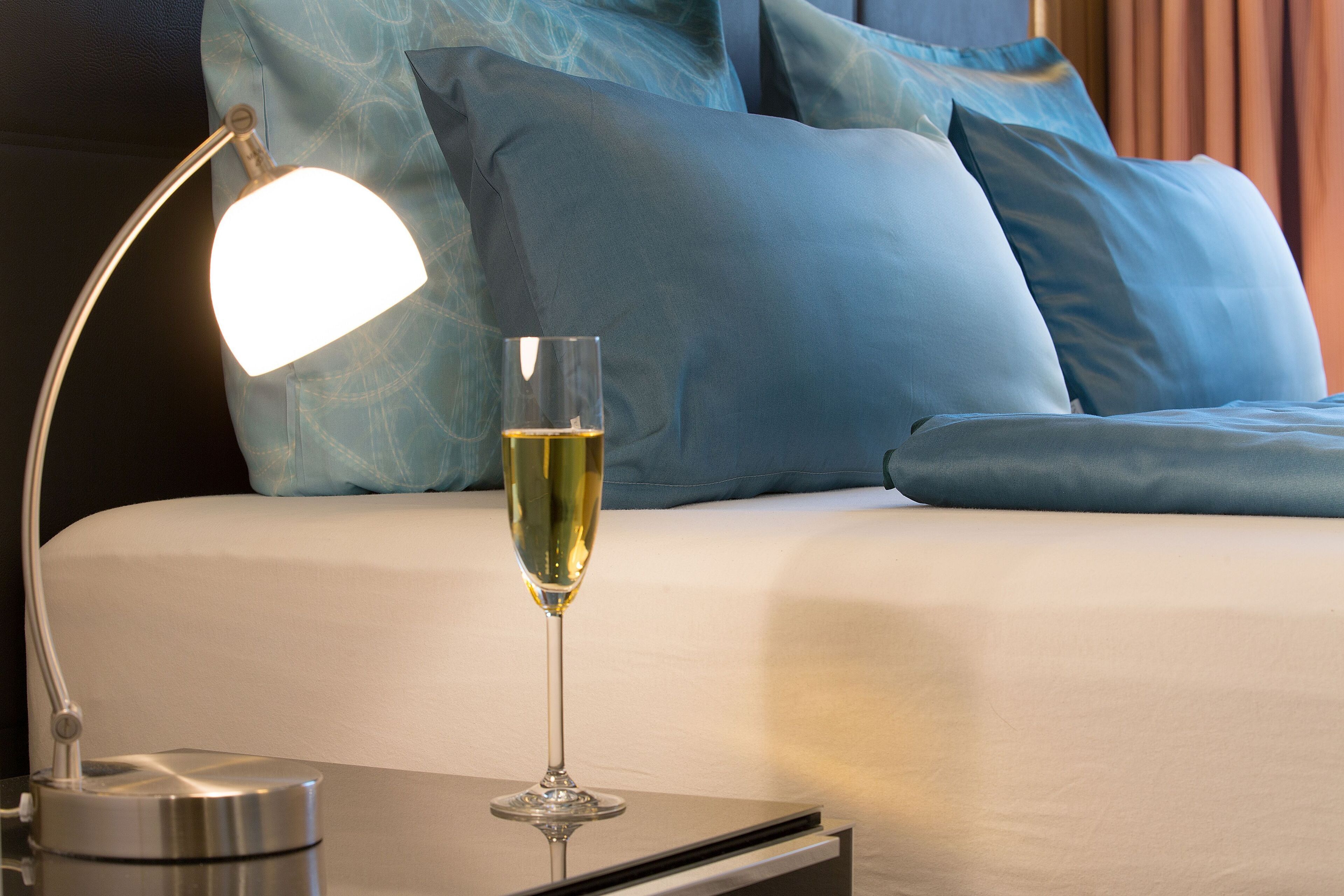 A neatly made bed with blue pillows, a bedside table with a lamp, and a glass of champagne.