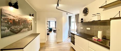 A modern kitchen with wooden flooring, white cabinetry, a black countertop, and a large cityscape painting on the wall.