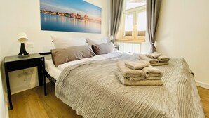 Un dormitorio con cama, almohadas, mesita de noche, lámpara, ventana con cortinas y un cuadro en la pared.
