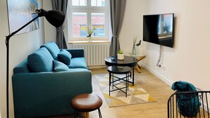 Una sala moderna con un sofá azul, una mesita redonda, un televisor y una ventana con cortinas.