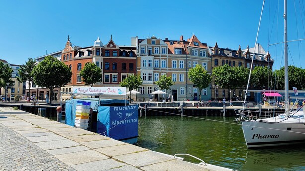 Una orilla con una hilera de edificios coloridos, un muelle con un velero llamado 'Pharoa', y un letrero que dice 'Frischfisch und Räucherverfisch'.