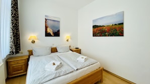 Una cama bien tendida con dos almohadas, una mesita de noche de madera, una lámpara y dos cuadros en la pared que muestran paisajes.