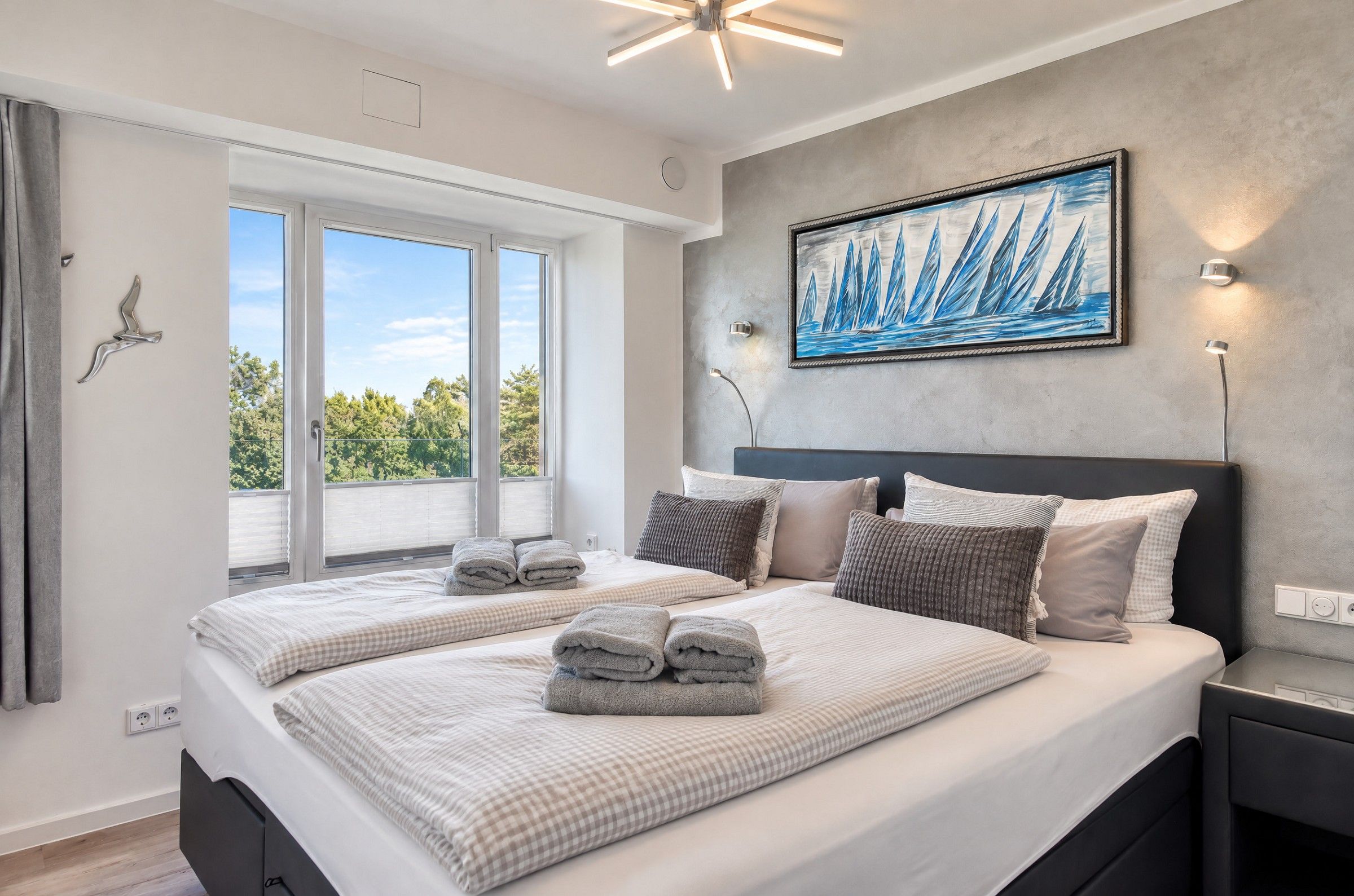 A modern bedroom with a large bed, a painting, and a view of greenery through the window.