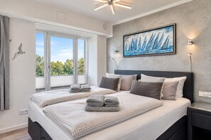 A modern bedroom with a large bed, a painting, and a view of greenery through the window.