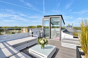 A rooftop terrace with a glass-enclosed seating area, a table with a vase of flowers, and a view of the surrounding landscape.