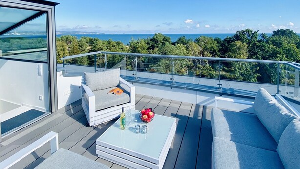 A modern outdoor seating area with a glass table, cushioned chairs, and a clear view of the ocean and trees.