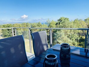 En balkon med et bord, to stole og udsigt til et skovområde og et fjerntliggende landskab.