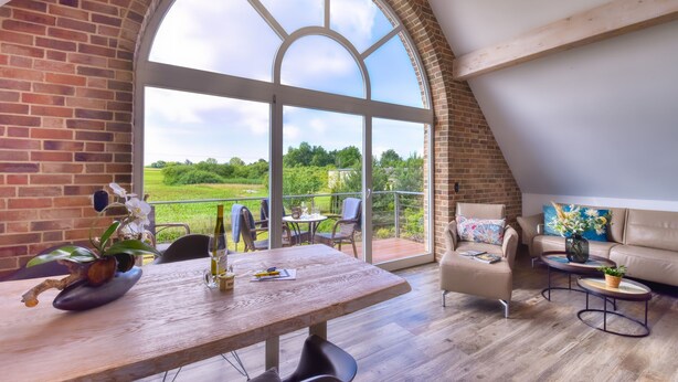 Un loft espacioso con un gran ventanal arqueado, una mesa de comedor de madera y una zona de estar acogedora con vistas a un paisaje verde.