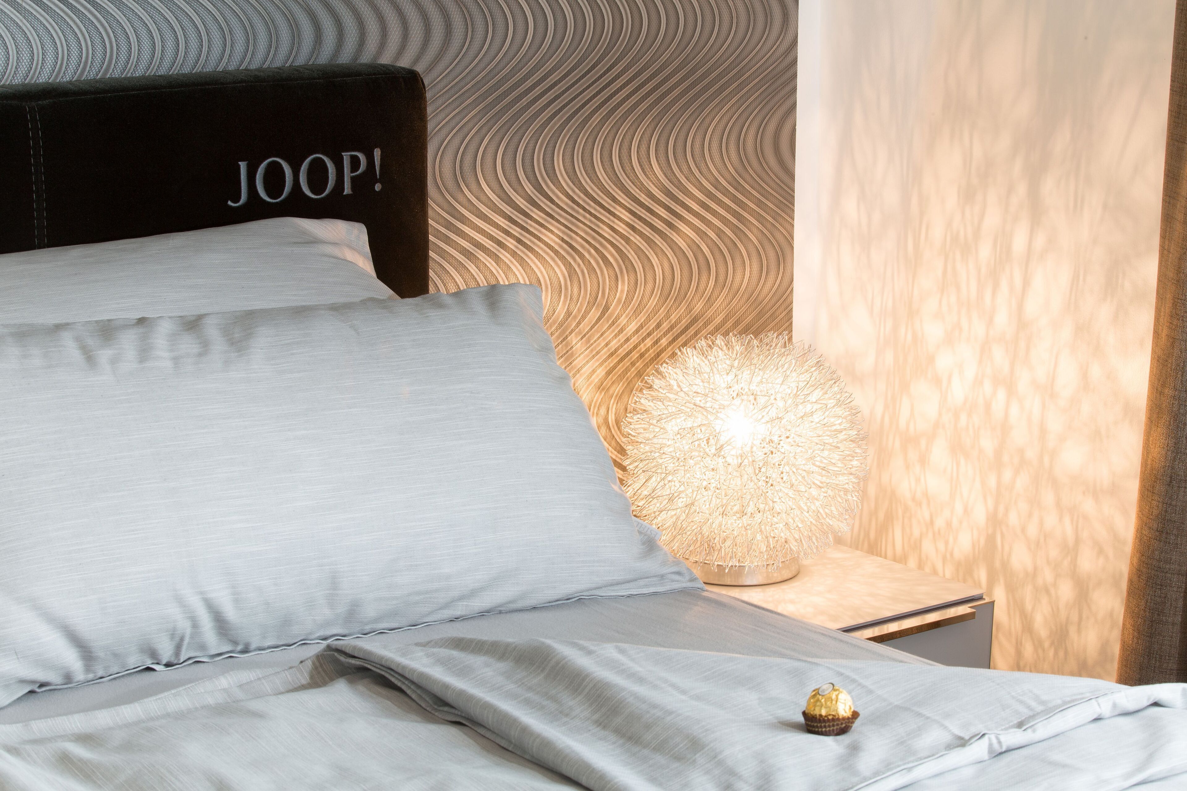 Un lit bien fait, avec une literie de couleur gris clair et un petit gâteau décoratif posé à côté.